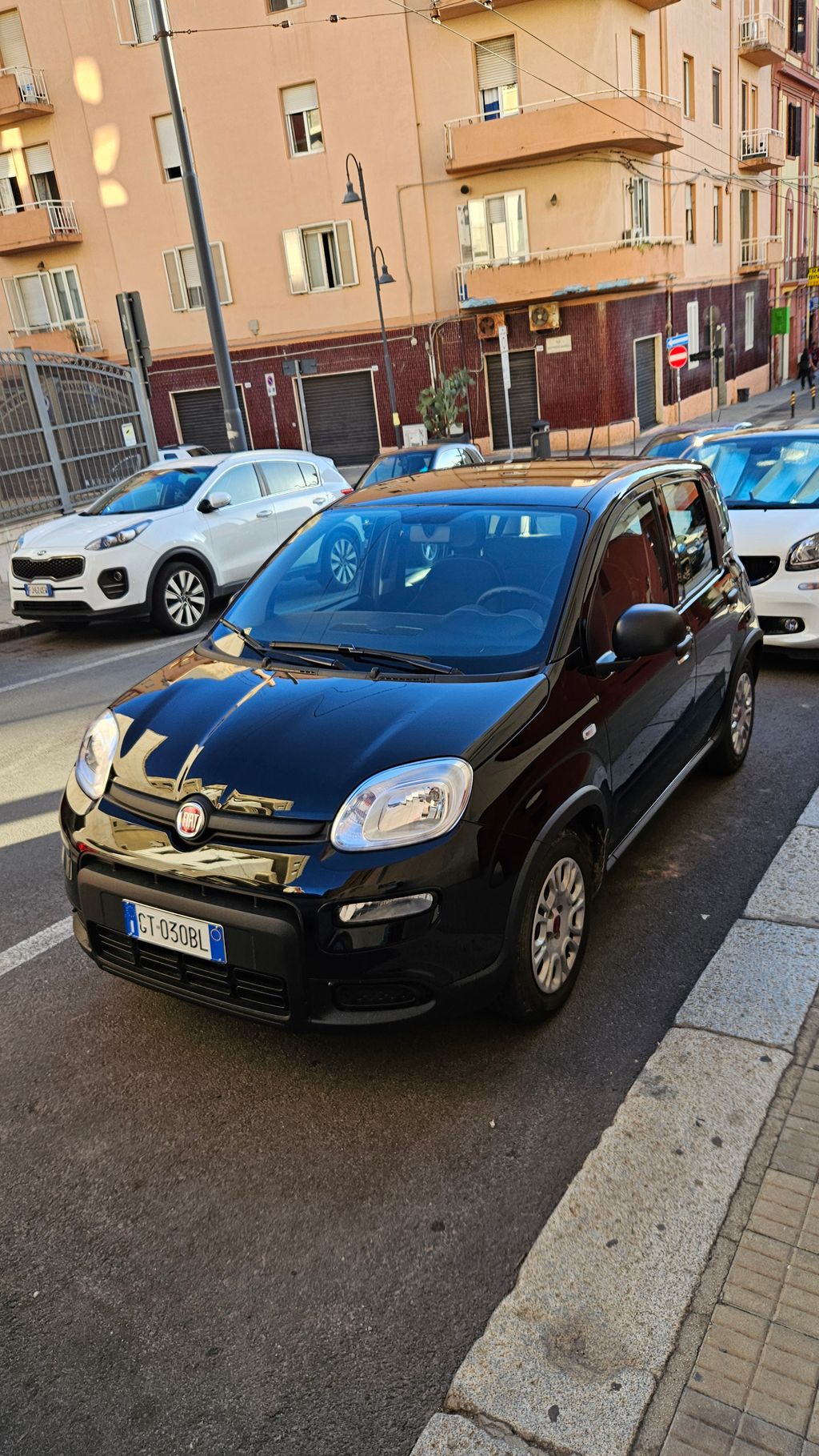 Schwarzer Fiat Panda auf einer Straße in einer städtischen Wohngegend mit weiteren parkenden Autos und einem mehrstöckigen Gebäude im Hintergrund.