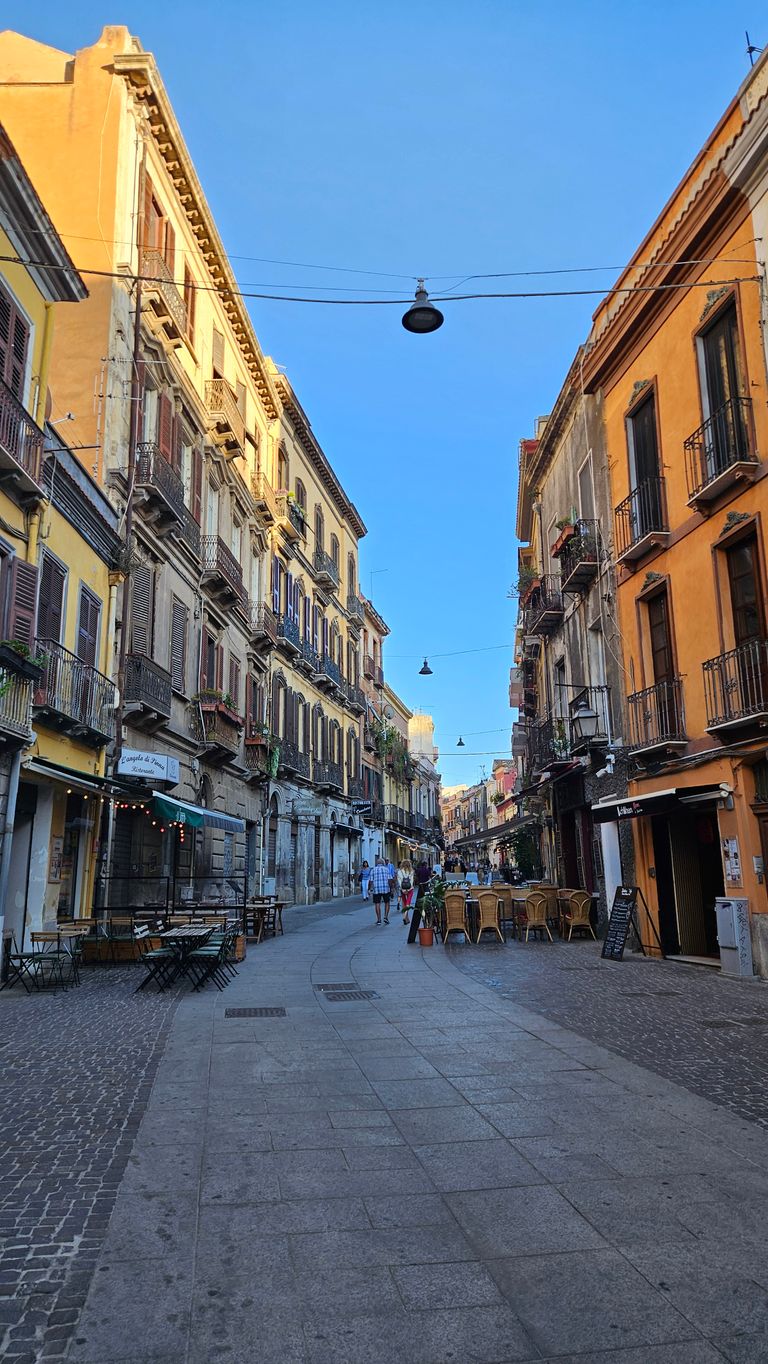 Eine gepflasterte Straße in einer belebten Altstadt mit mehrstöckigen Gebäuden und Außensitzbereichen von Restaurants bei klarem Himmel.