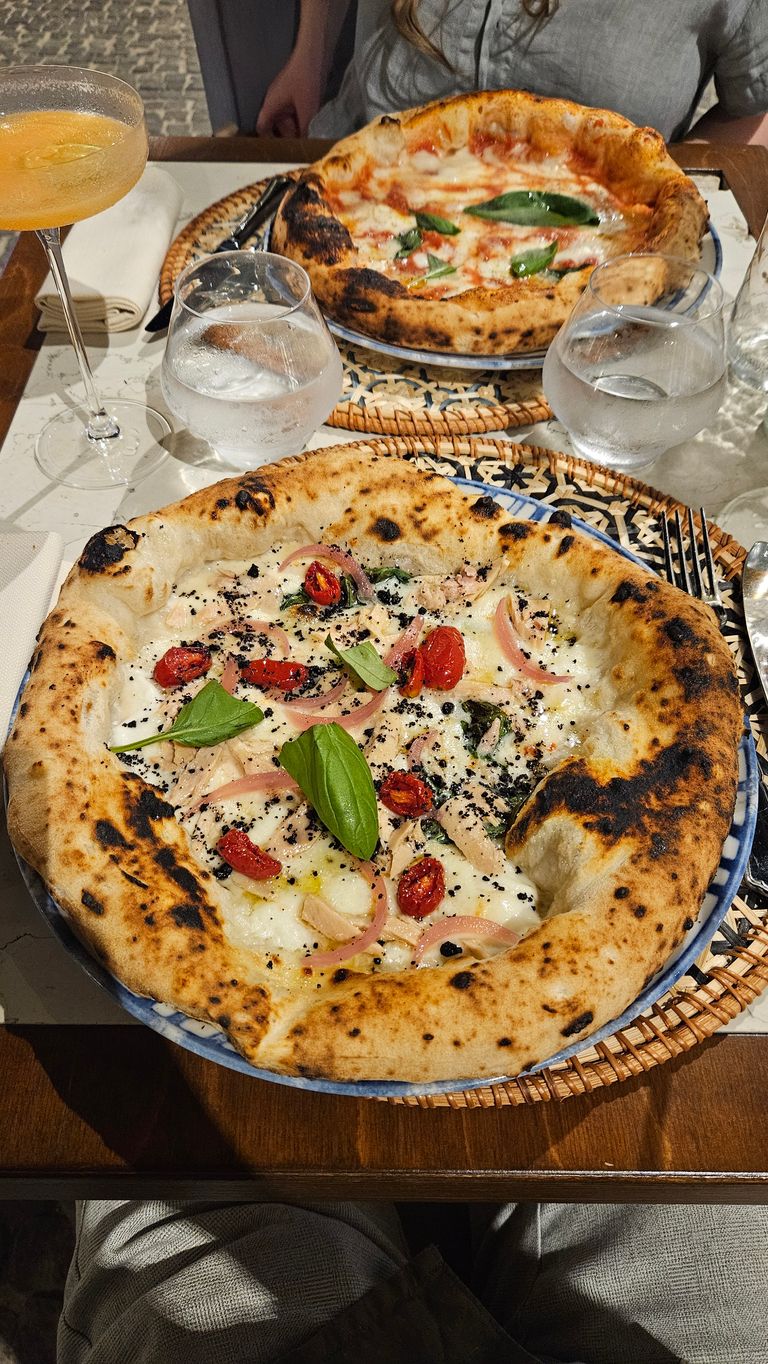 Zwei neapolitanische Pizzen auf einem Holztisch, umgeben von Gläsern mit Wasser und einem Cocktail, in einem gemütlichen Restaurant.