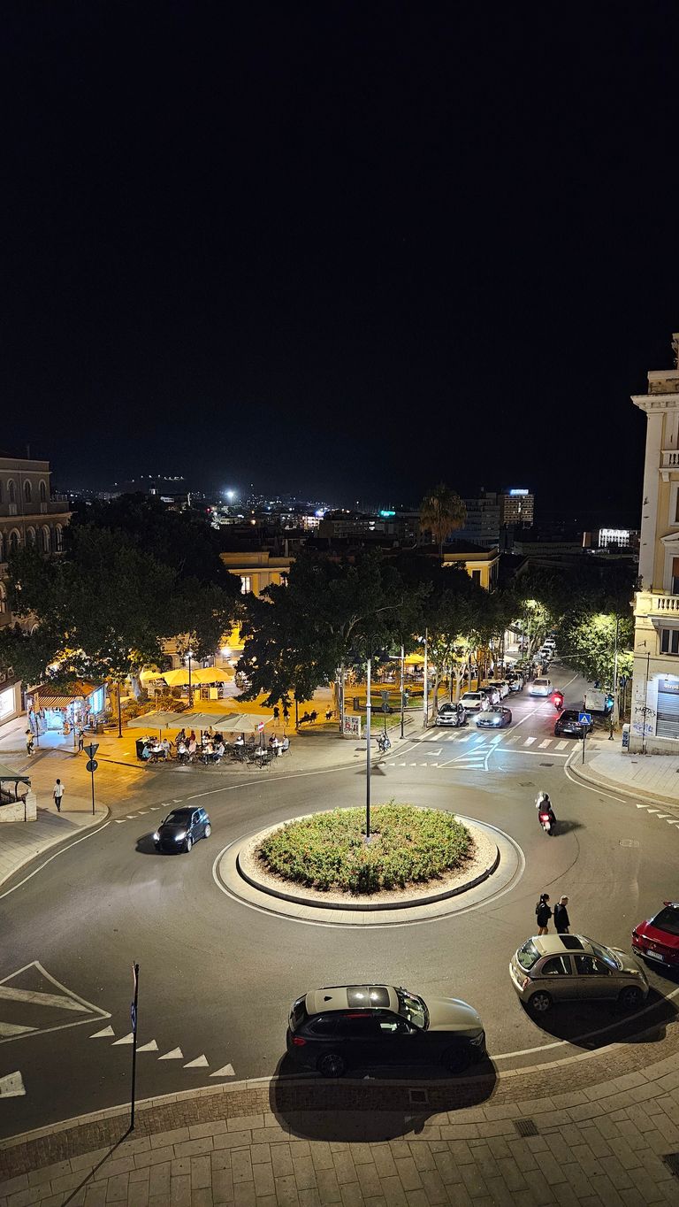 Blick auf einen beleuchteten Kreisverkehr mit einer bepflanzten Mittelinsel in einer nächtlichen Stadtumgebung, umgeben von Autos, Fußgängern und einem Café mit Sitzmöglichkeiten im Freien.
