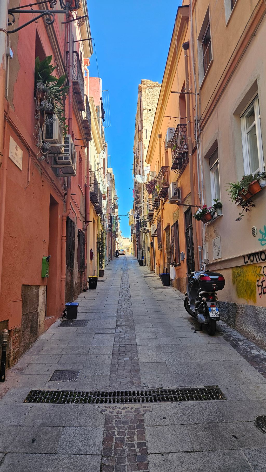 Enge, gepflasterte Gasse zwischen farbigen Gebäuden mit Balkonen und Fenstern, links rote und rechts beige Fassaden, bei klarem Himmel.
