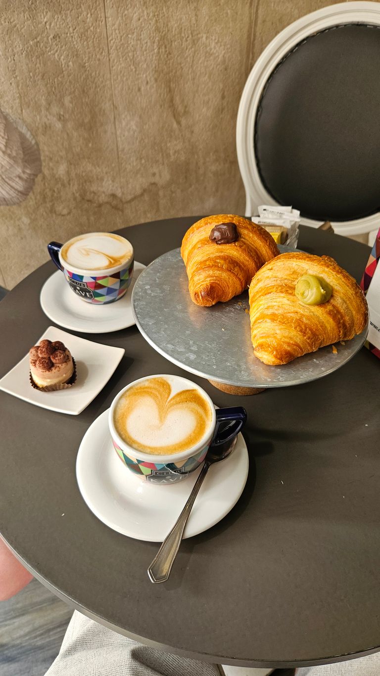 Zwei bunte Cappuccino-Tassen auf weißen Untertellern mit Milchschaum, zwei Croissants mit unterschiedlicher Füllung auf einem Metallteller und ein kleines Schokoladen-Mousse-Dessert auf einem quadratischen weißen Teller auf einem runden grauen Tisch.