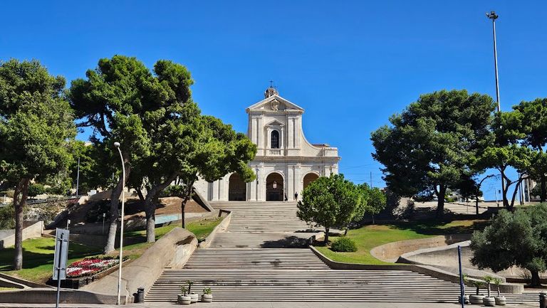 Treppenaufgang zu einer hellen Kirche mit klassizistischer Fassade, umgeben von grünen Bäumen und unter blauem Himmel.