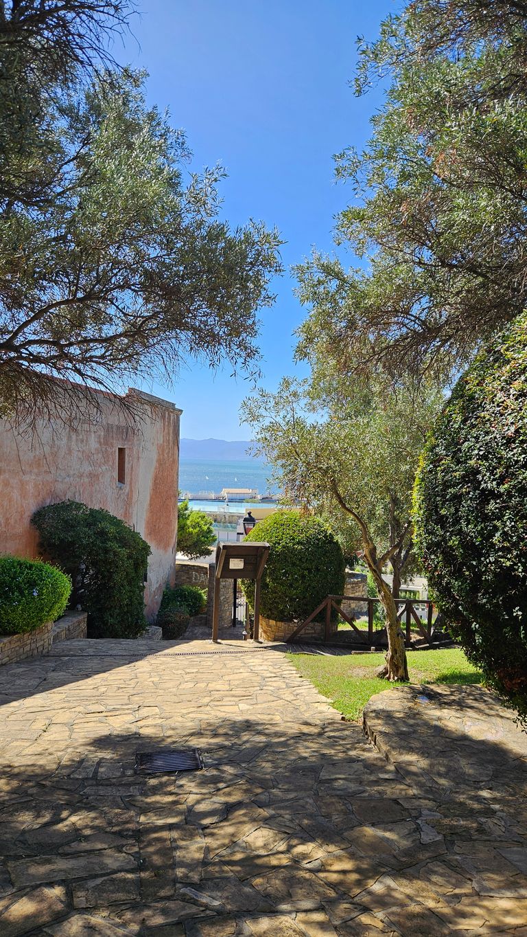 Ein schattiger, gepflasterter Weg führt zwischen grünen Büschen und einem alten Gebäude mit einer Meerblick-Kulisse im Hintergrund unter klarem blauem Himmel.