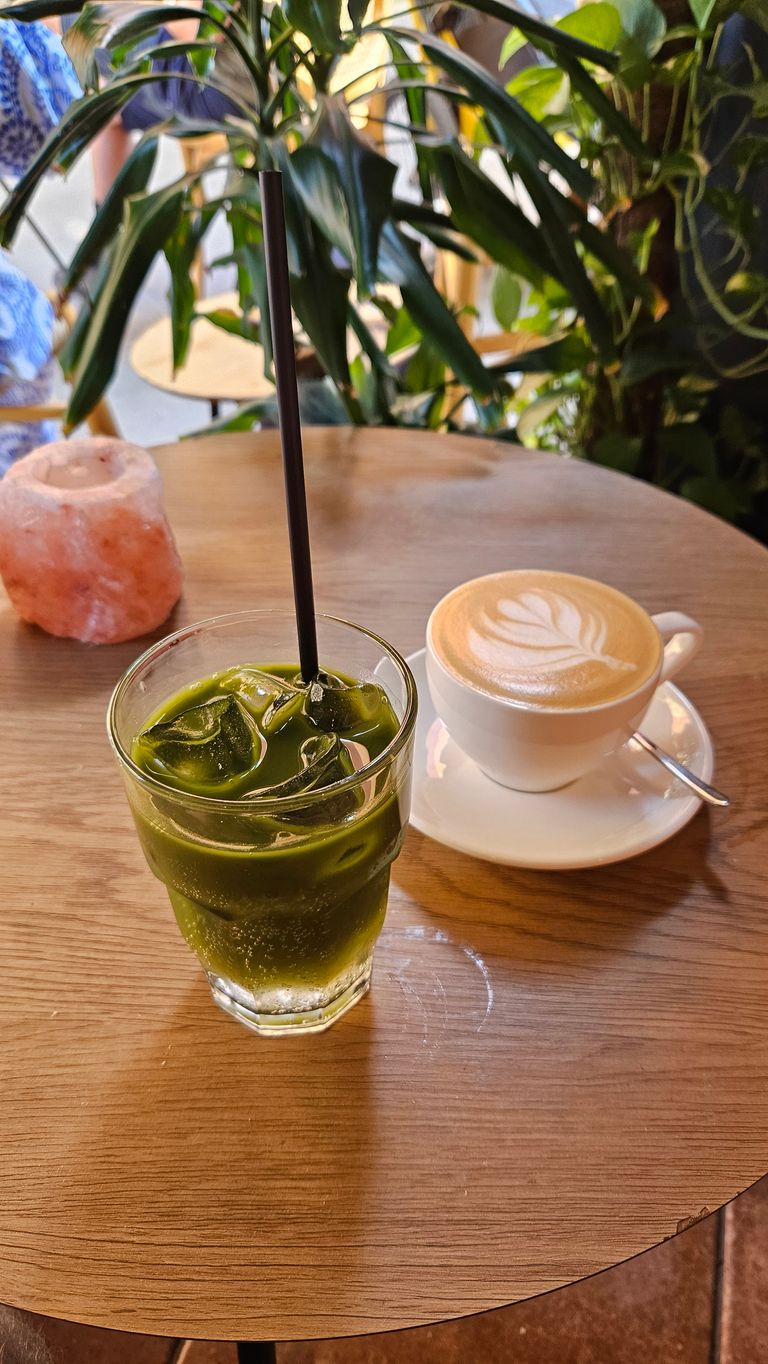 Ein Glas mit grünem Eistee und einem schwarzen Strohhalm steht auf einem runden Holztisch neben einer weißen Tasse mit Latte Art und Untertasse, umgeben von grünen Pflanzen in einem Café.