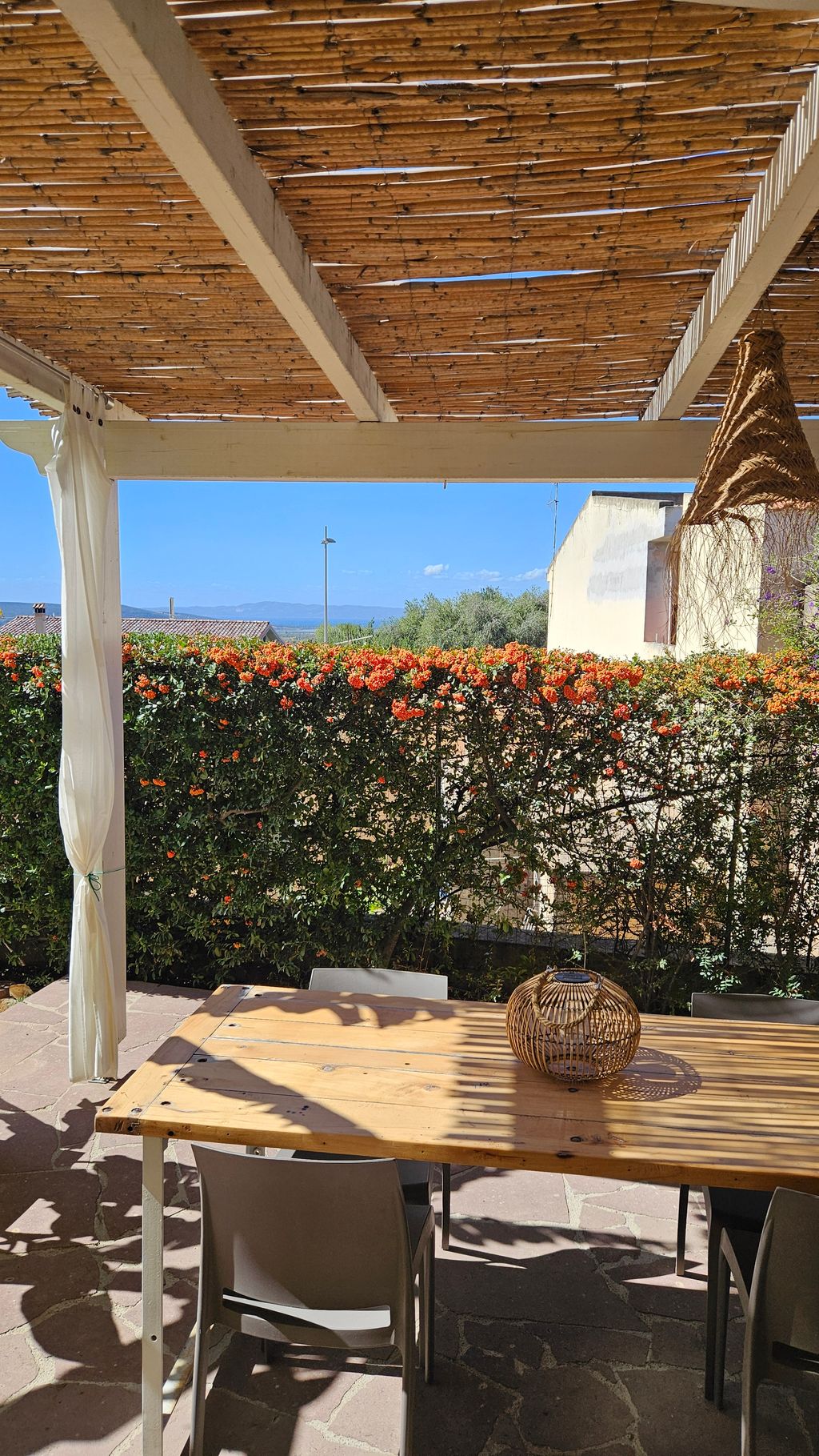 Überdachte Terrasse mit Holztisch, mehreren Stühlen und einer Laterne aus Weide, umgeben von grünen Hecken mit orangefarbenen Blüten, bei sonnigem Wetter und blauem Himmel.