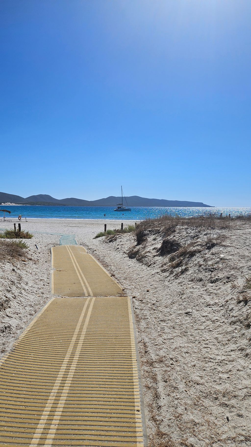 Ein langer gelber Strandweg aus Kunststoffmatten, der durch weiße Sanddünen zum blauen Meer mit einem Anker-Katamaran und Bergen im Hintergrund führt, unter klarem blauem Himmel.
