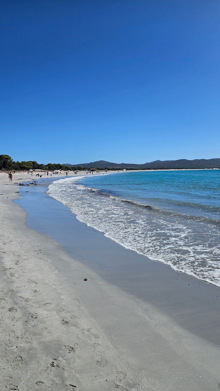 Weitläufiger Sandstrand mit sanften Wellen und blauem Himmel, im Hintergrund sind Menschen am Strand und Berge sichtbar.