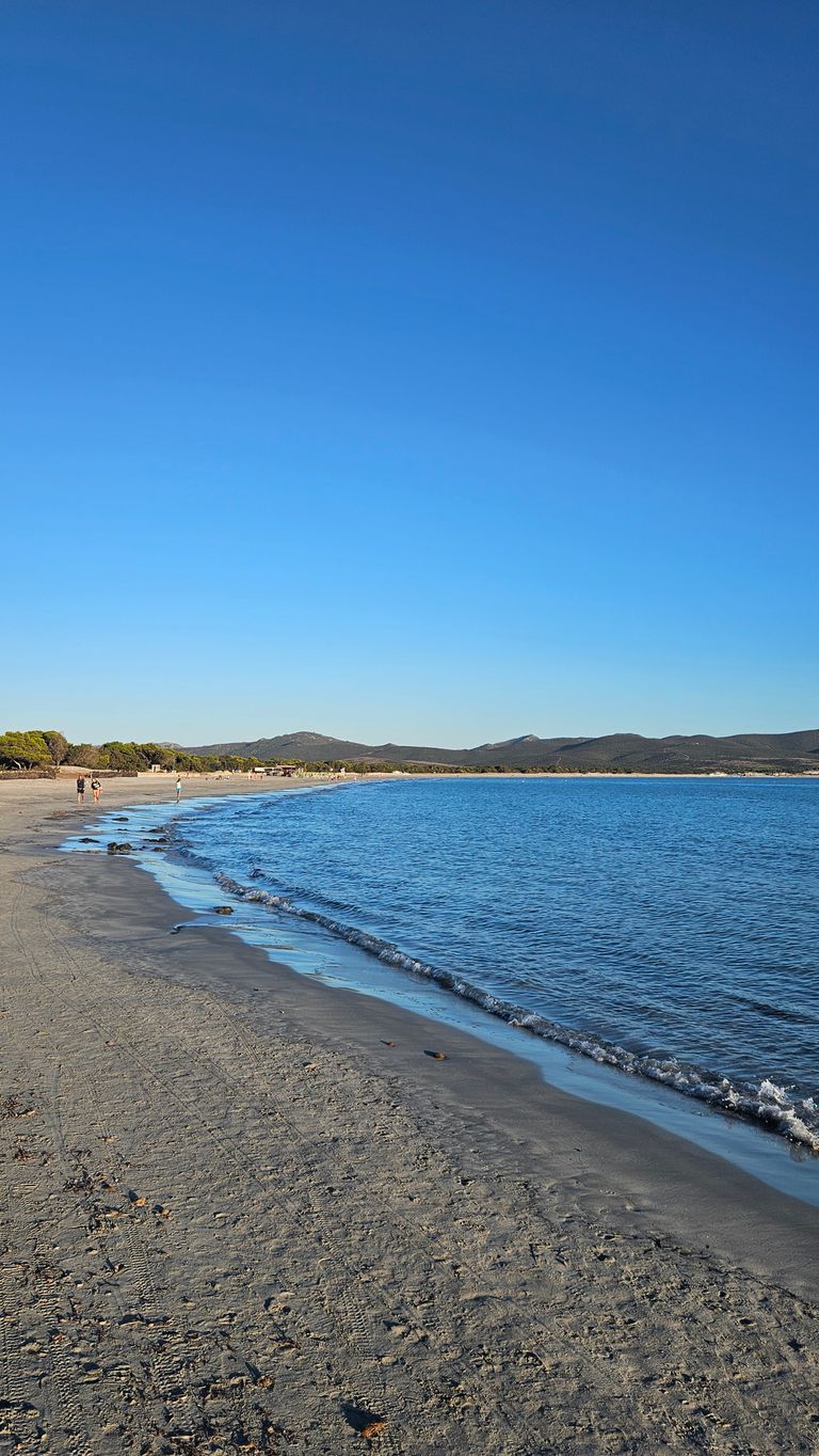 Strand mit nassem Sand und klarem blauen Meer unter einem wolkenlosen, tiefblauen Himmel. Am Strand sind in der Ferne einige Personen zu sehen, und im Hintergrund sind hügelige Landschaften mit Bäumen erkennbar.