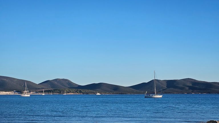 Segelboote auf ruhigem blauen Meer vor einer hügeligen Küstenlandschaft unter klarem blauen Himmel.