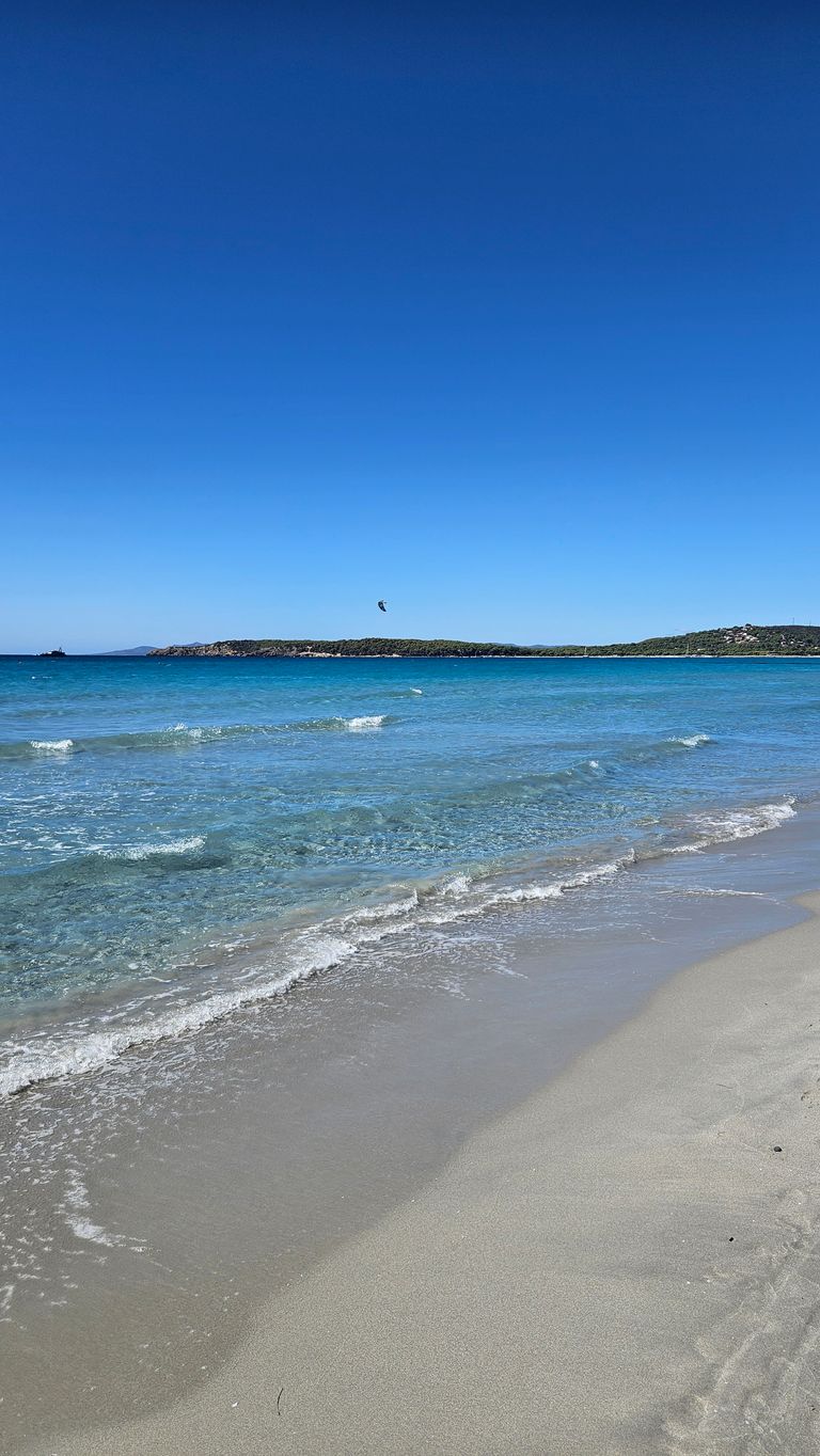 Klares, ruhiges Meer mit kleinen Wellen an einem Sandstrand, im Hintergrund bewaldete Inseln unter blauem Himmel.