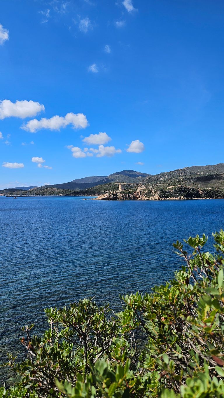 Blauer Himmel mit weißen Wolken über einer Küstenlandschaft mit klarem, blauem Wasser und grünen Büschen im Vordergrund. Im Hintergrund sind Hügel und ein runder Steinturm auf einer Felseninsel zu sehen.