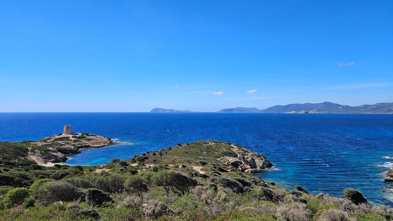 Küstenlandschaft mit grüner Vegetation, einem kleinen Turm auf einer Landzunge und klarem blauem Meer unter hellem Himmel.