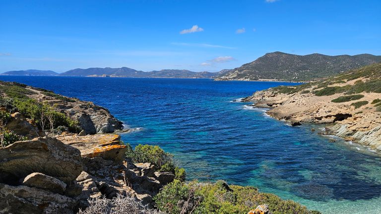 Küstenlandschaft mit klarem blauem Meer, felsigen Ufern und bewachsenen Hügeln bei sonnigem Himmel.