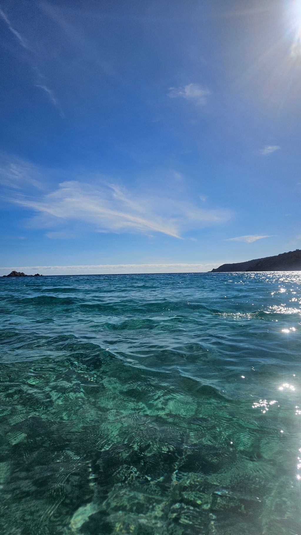 Klares Meer mit sichtbarem Meeresboden unter blauem Himmel und einem bewachsenen Hügel am Horizont rechts.