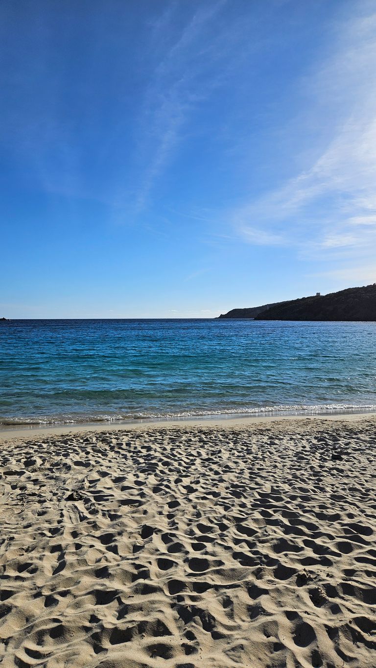 Sandstrand mit Wasser und einem bewaldeten Kap im Hintergrund unter einem blauen Himmel mit wenigen Wolken.