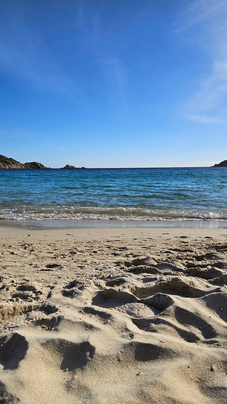 Sandstrand im Vordergrund mit Muscheln, dahinter ruhiges Meer und im Hintergrund Felsen unter klarem blauem Himmel.