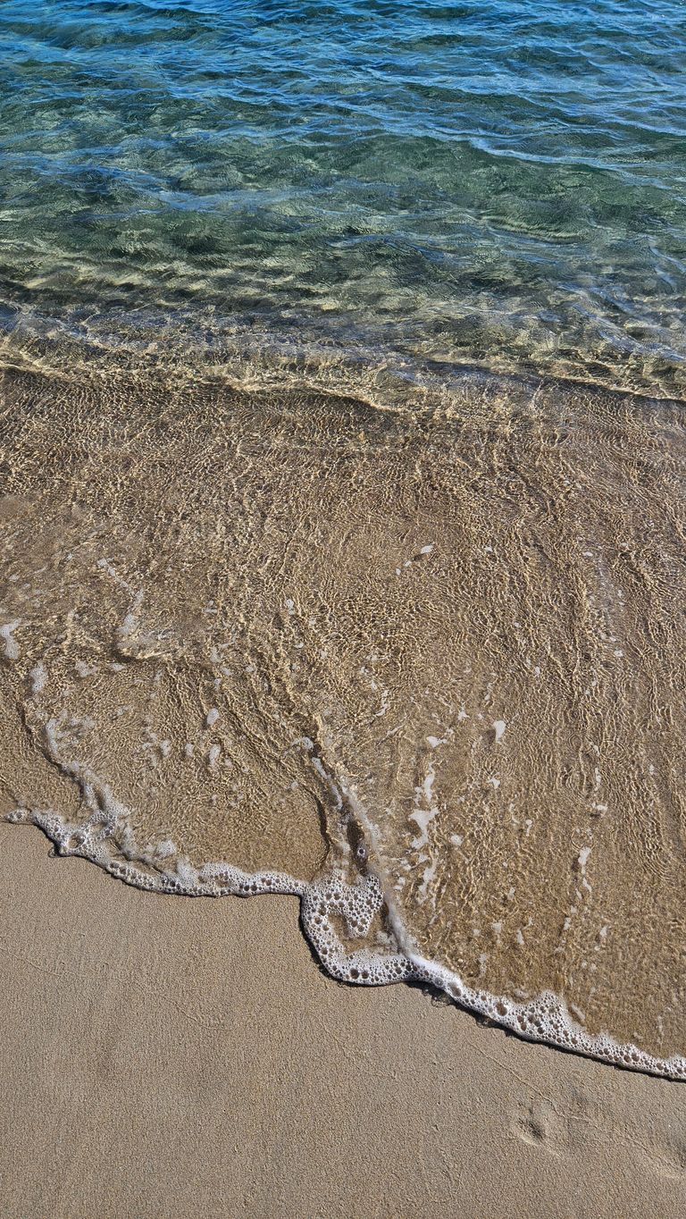 Klares Wasser brandet sanft auf einen Sandstrand mit sichtbaren Schaumkronen.