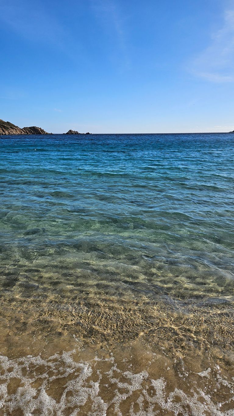 Klares, ruhiges Meer mit transparentem Wasser und leichtem Wellengang am Sandstrand unter blauem Himmel mit ein paar Felsen am Horizont.