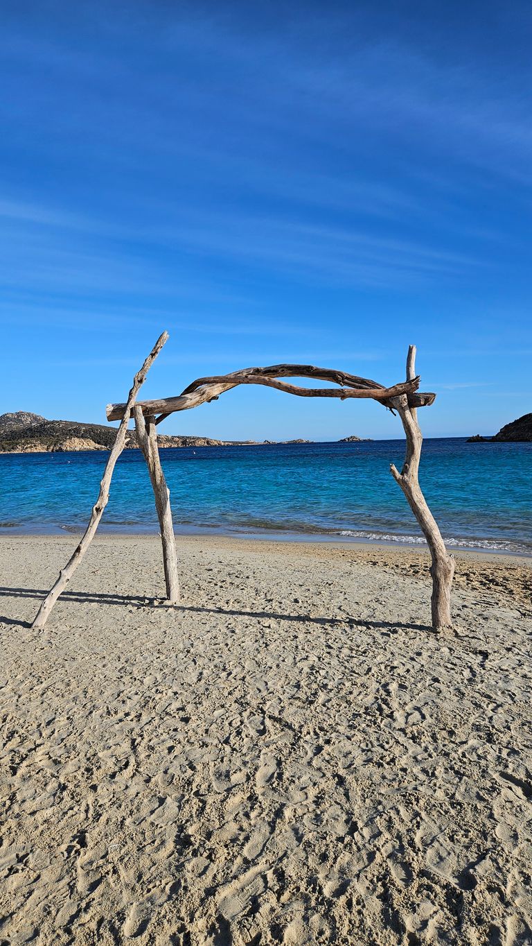Ein einfaches Gestell aus Treibholz steht auf einem Sandstrand vor einer ruhigen, blauen Meeresküste unter hellem Himmel.