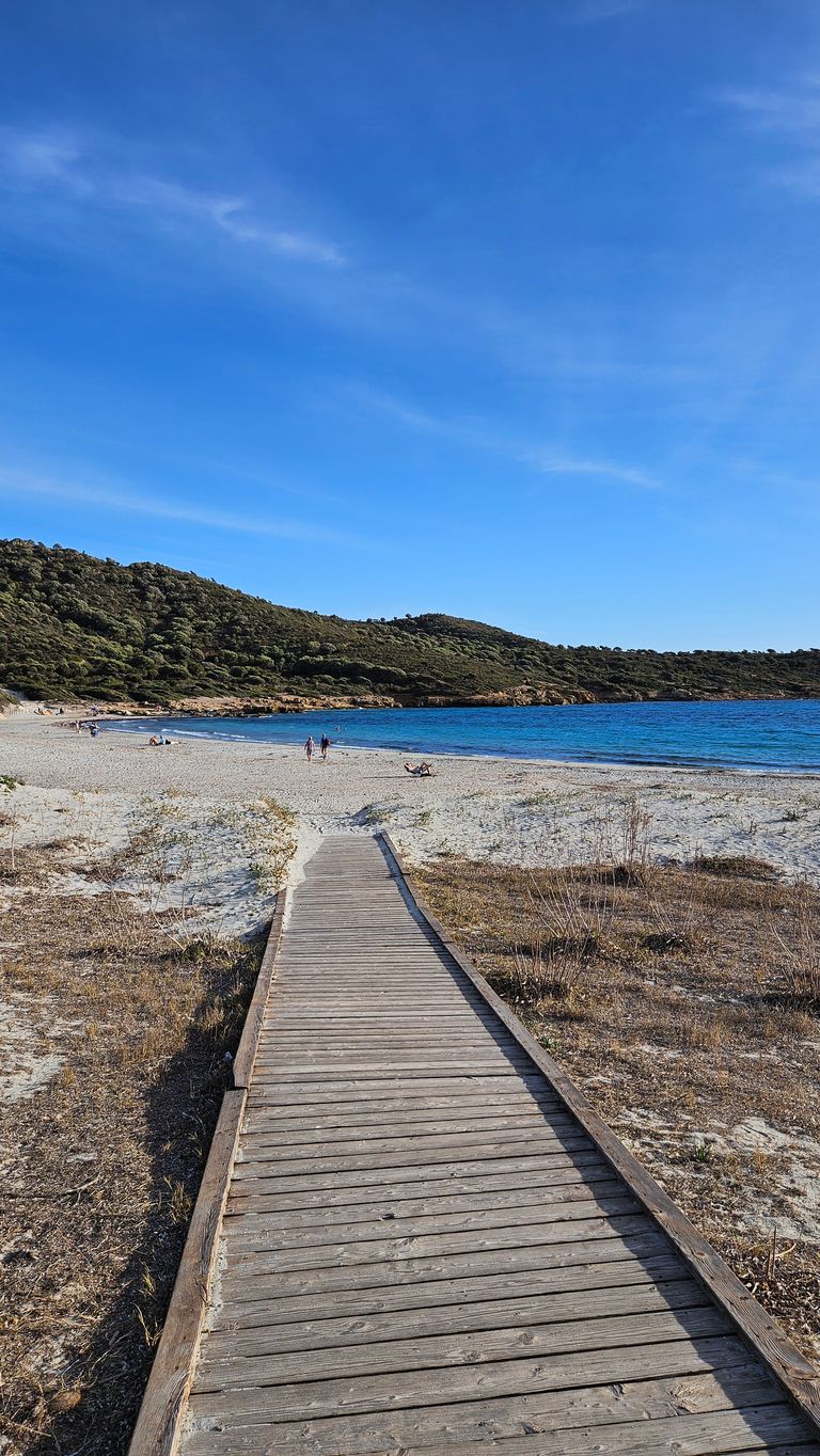 Ein Holzsteg führt über trockene Vegetation zu einem Sandstrand mit einigen wenigen Menschen, davor ist das blaue Meer und dahinter bewaldete Hügel unter blauem Himmel zu sehen.