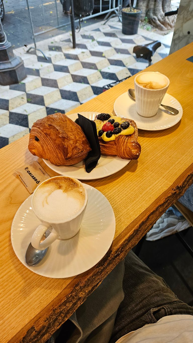 Zwei Tassen Cappuccino und ein Teller mit zwei Gebäckstücken, darunter ein mit Beeren belegtes Croissant, auf einem Holzthekenplatz an einem Café-Fenster mit Blick auf den gepflasterten Gehweg.