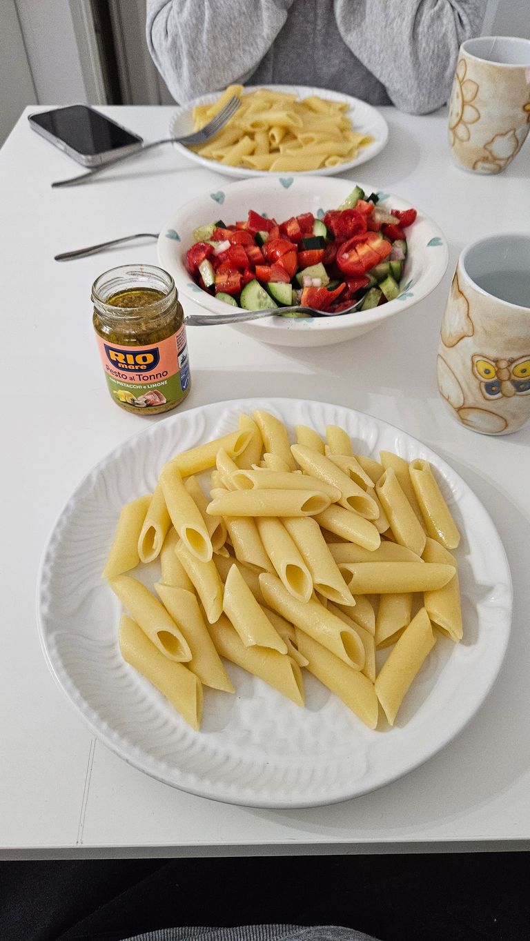 Teller mit Penne-Nudeln, eine Schüssel mit Gurken- und Paprikasalat, ein Glas Pesto al Tonno und zwei Tassen stehen auf einem weißen Tisch. Im Hintergrund sitzt eine Person mit grauer Kleidung, dahinter liegt ein Smartphone.