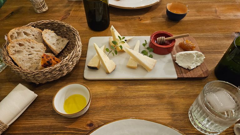 Ein Holztisch mit einem weißen Teller mit mehreren Käsekeilen, einem kleinen roten Gefäß mit Honiglöffel, Brot in einem Weidenkorb, einem kleinen Schälchen mit Olivenöl, einem Glas mit Eiswürfeln und mehreren Flaschen oder Gläsern.