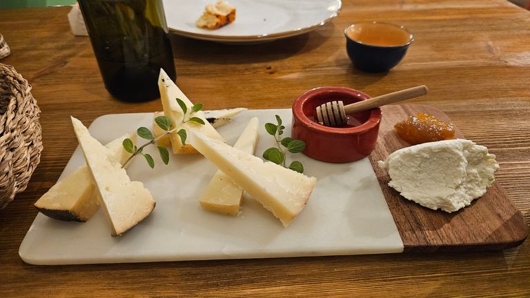 Auf einem rechteckigen Schneidebrett aus Marmor und Holz liegen mehrere dreieckige Käsestücke, ein Klecks Frischkäse, ein kleiner roter Tontopf mit Honiglöffel und etwas Marmelade. Das Brett steht auf einem Holztisch, im Hintergrund sind eine Flasche, eine Tasse und ein Teller zu sehen.