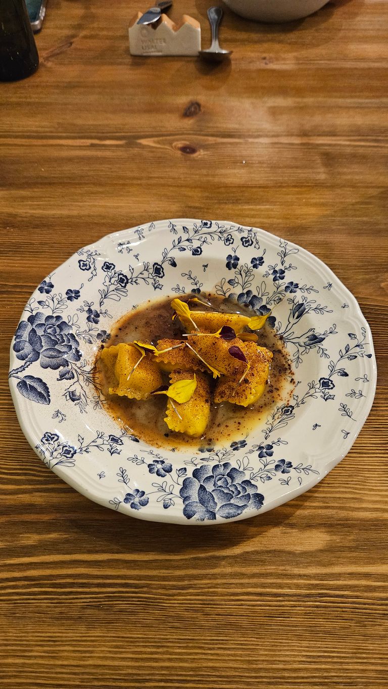 Gelbe Ravioli in einer braunen Sauce, garniert mit essbaren Blüten, serviert auf einem weißen Teller mit dekorativem blauen Blumenmuster auf einem Holztisch.