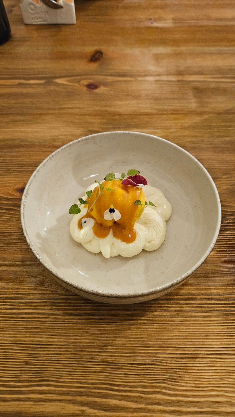 Ein Dessert in einer beigen Schale auf einem Holztisch bestehend aus einer Mousse mit oranger Sauce und essbaren Blüten und Kräutern garniert.