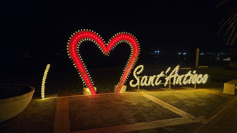 Illuminiertes Leuchtschild mit Herz und der Aufschrift "Sant'Antioco" an einem dunklen Abend auf einem gepflasterten Platz.