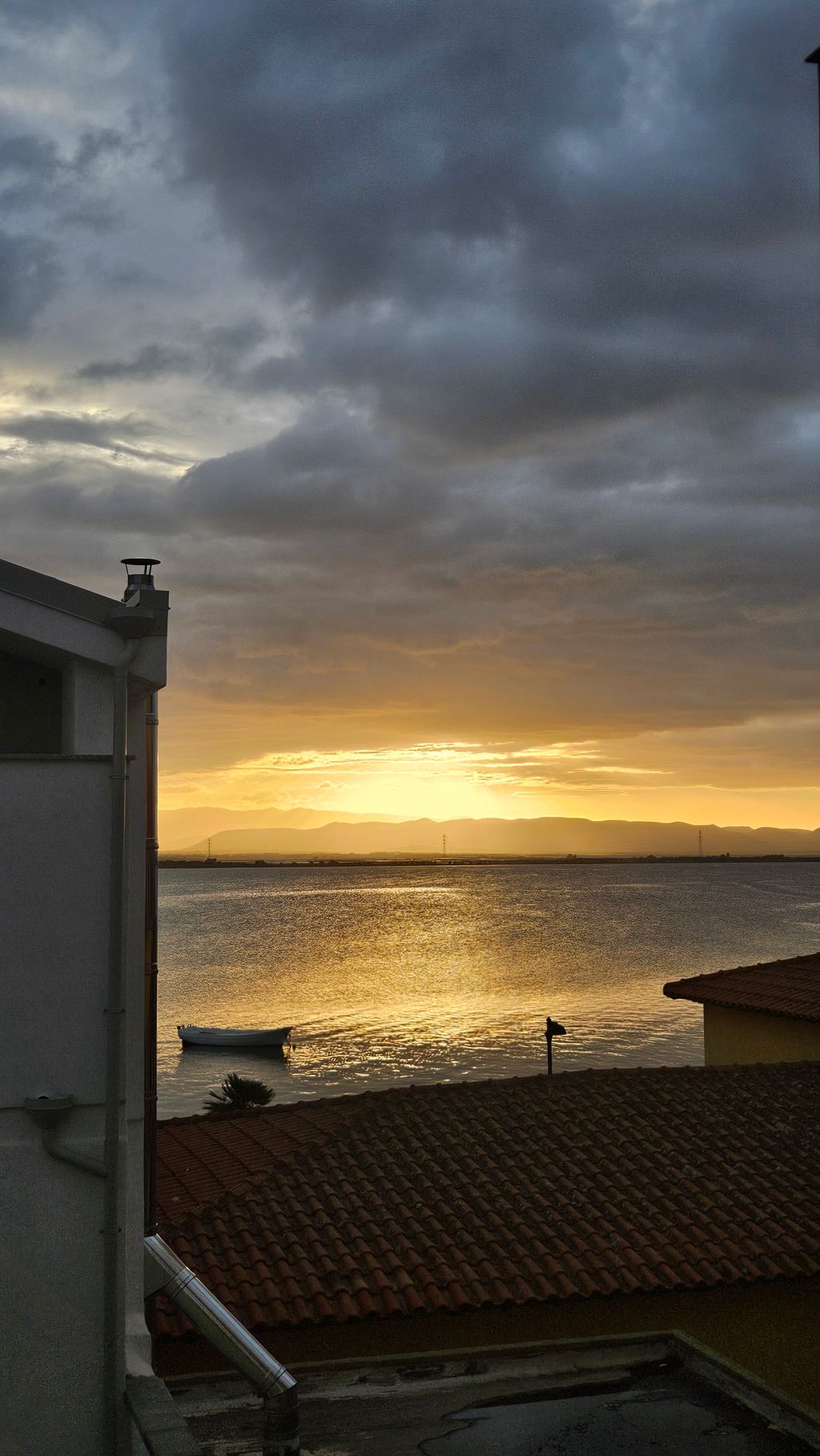 Sonnenaufgang über einem ruhigen See mit einem kleinen Boot und dunklen Wolken am Himmel, im Vordergrund ein rotes Ziegeldach und Gebäudeecken.
