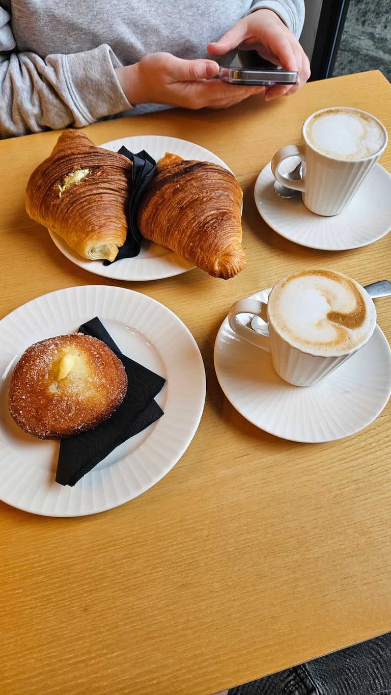 Zwei Tassen Cappuccino und drei Gebäckstücke auf weißen Tellern auf einem Holztisch, eine Person im grauen Pullover hält ein Smartphone.
