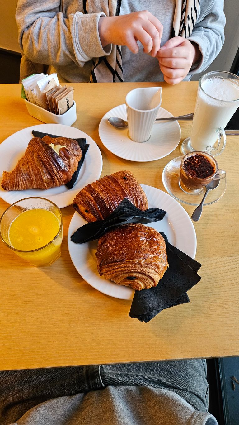Frühstückstisch mit Croissants, Café, Orangensaft und Milchgetränk, mit einer Person im Hintergrund, die ihre Hände hält.