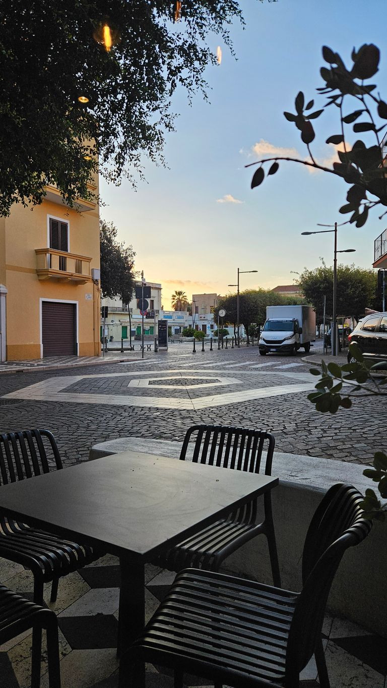 Blick von einem Außentisch mit schwarzen Stühlen auf einen gepflasterten Platz mit dekorativem Muster, umgeben von Gebäuden und Bäumen bei Sonnenaufgang oder Sonnenuntergang.