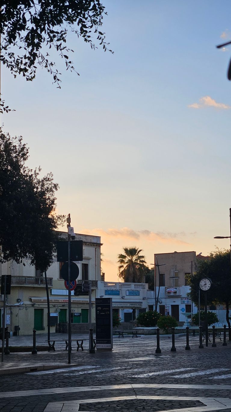 Ein ruhiger, leerer Platz in einer Stadt bei Sonnenaufgang mit Gebäuden, Bäumen und einer großen Uhr auf einer Straßenecke.