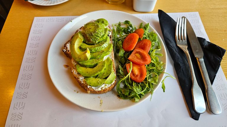 Ein Teller mit einem Avocado-Brot und einem Salat aus Rucola und Tomatenscheiben auf einem Tisch mit Besteck auf schwarzer Serviette.