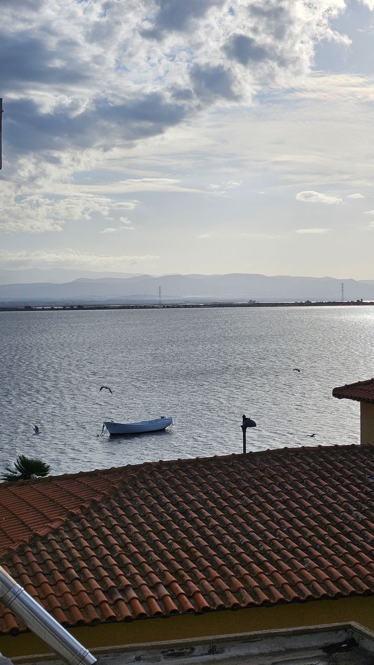 Blick über ein ruhiges Gewässer mit einem kleinen Boot, umgeben von Dächern mit roten Ziegeln und sich im Hintergrund erstreckenden Bergen unter einem bewölkten Himmel.