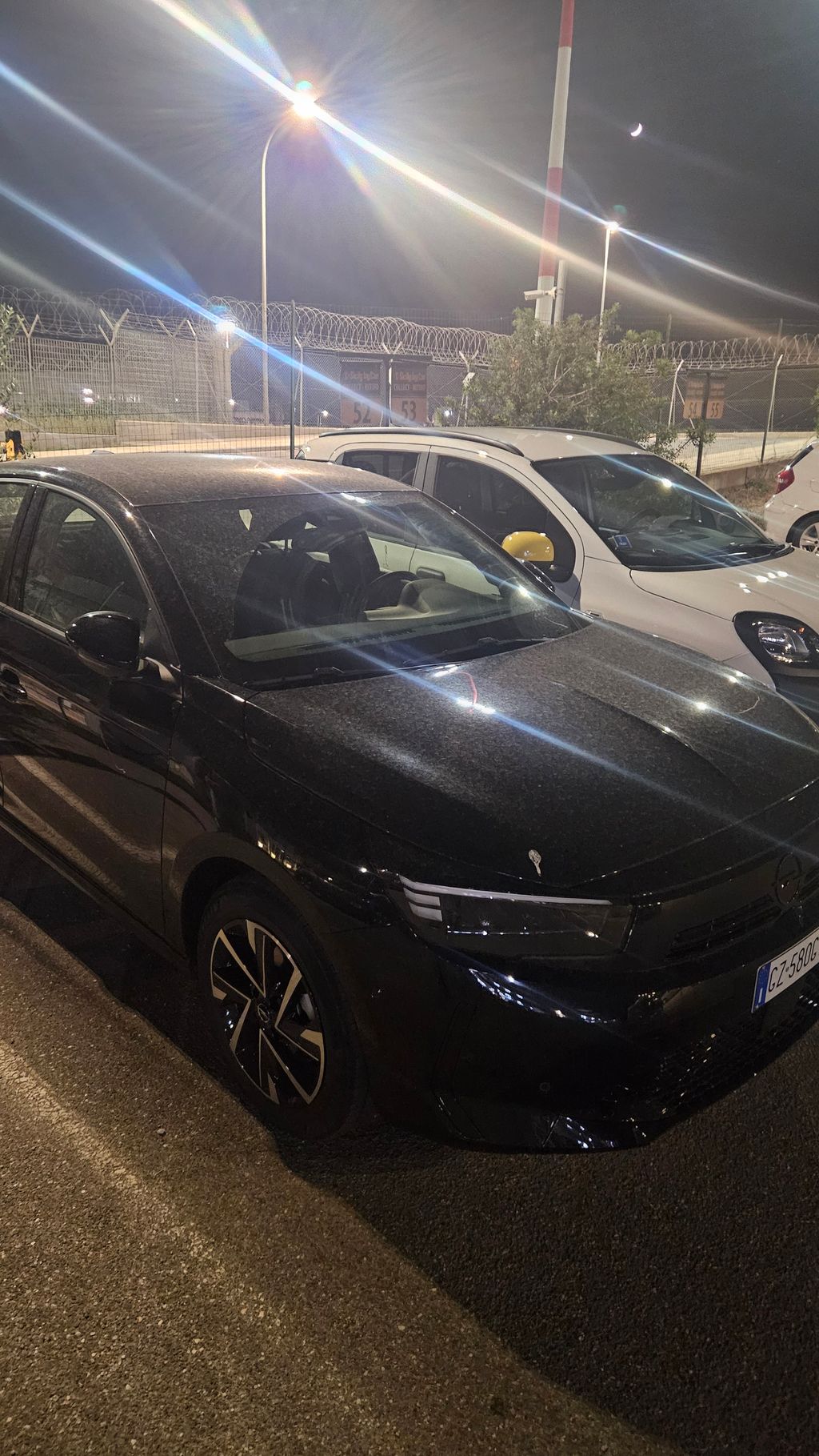Zwei geparkte Autos, davon ein schwarzes und ein weißes, nachts auf einem Parkplatz mit hell erleuchteten Straßenlaternen und einem Zaun mit Stacheldraht im Hintergrund.