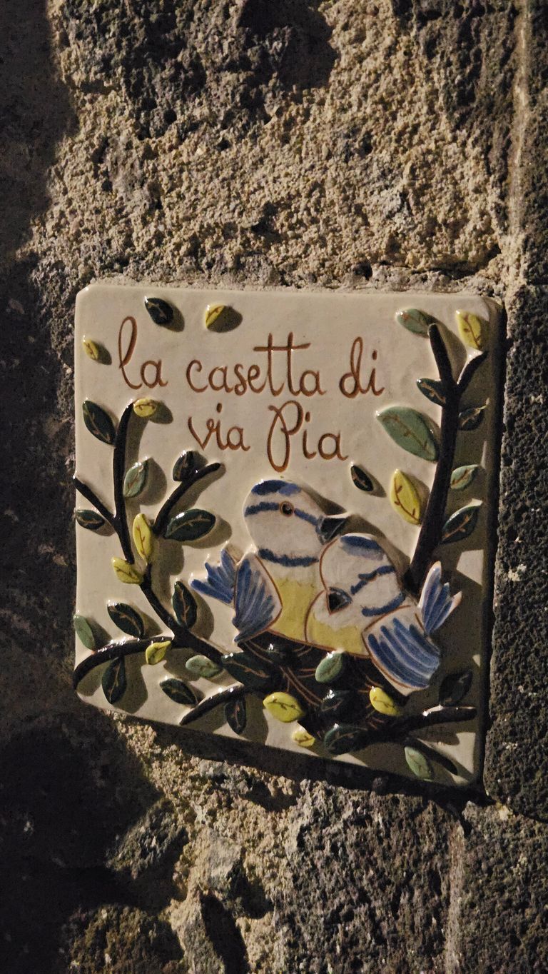 Keramikschild an einer rauen Steinwand mit der Aufschrift 'la casetta di via Pia' und bemalten Vögeln und Blättern darauf.