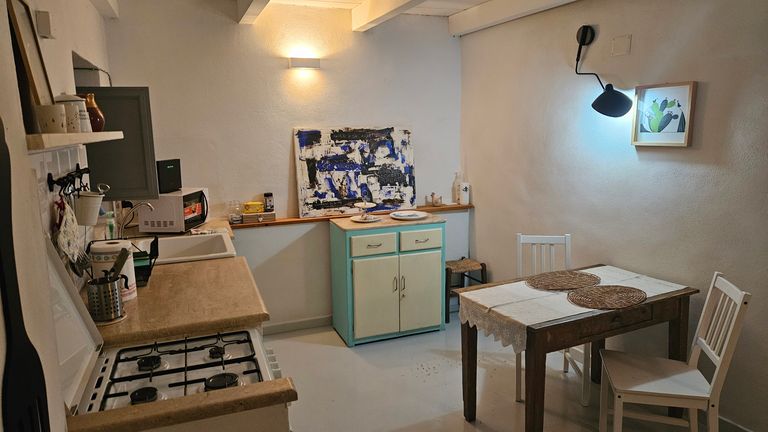 Kleine Küche mit Holztisch, zwei weißen Stühlen, einem alten Gasherd, einer Spüle und einem Unterbauschrank mit türkisfarbenen Akzenten. An der Wand hängt ein abstraktes Gemälde und eine beleuchtete Lampe über einem Kaktus-Bild.