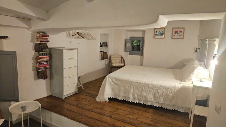 Schlafzimmer mit Doppelbett mit weißer Bettdecke, einem weißen Nachttisch mit Lampe, einem weißen Kommodenschrank, einem Stuhl mit Kissen und Büchern an der Wand in einem Raum mit Holzboden und weiß gestrichenen Wänden.