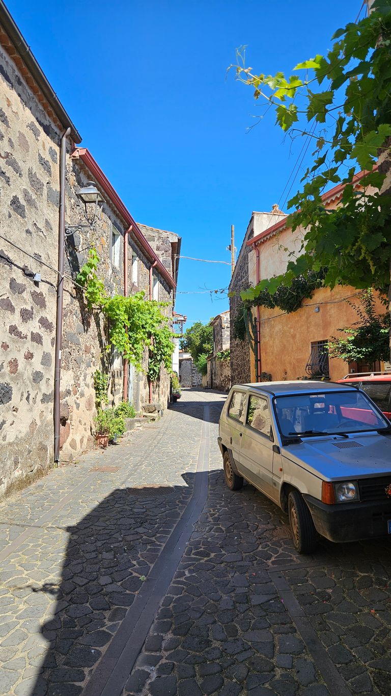 Eine enge Gasse mit Kopfsteinpflaster zwischen alten Stein- und verputzten Gebäuden, einem alten Auto rechts und grünen Pflanzen, unter einem klaren blauen Himmel.