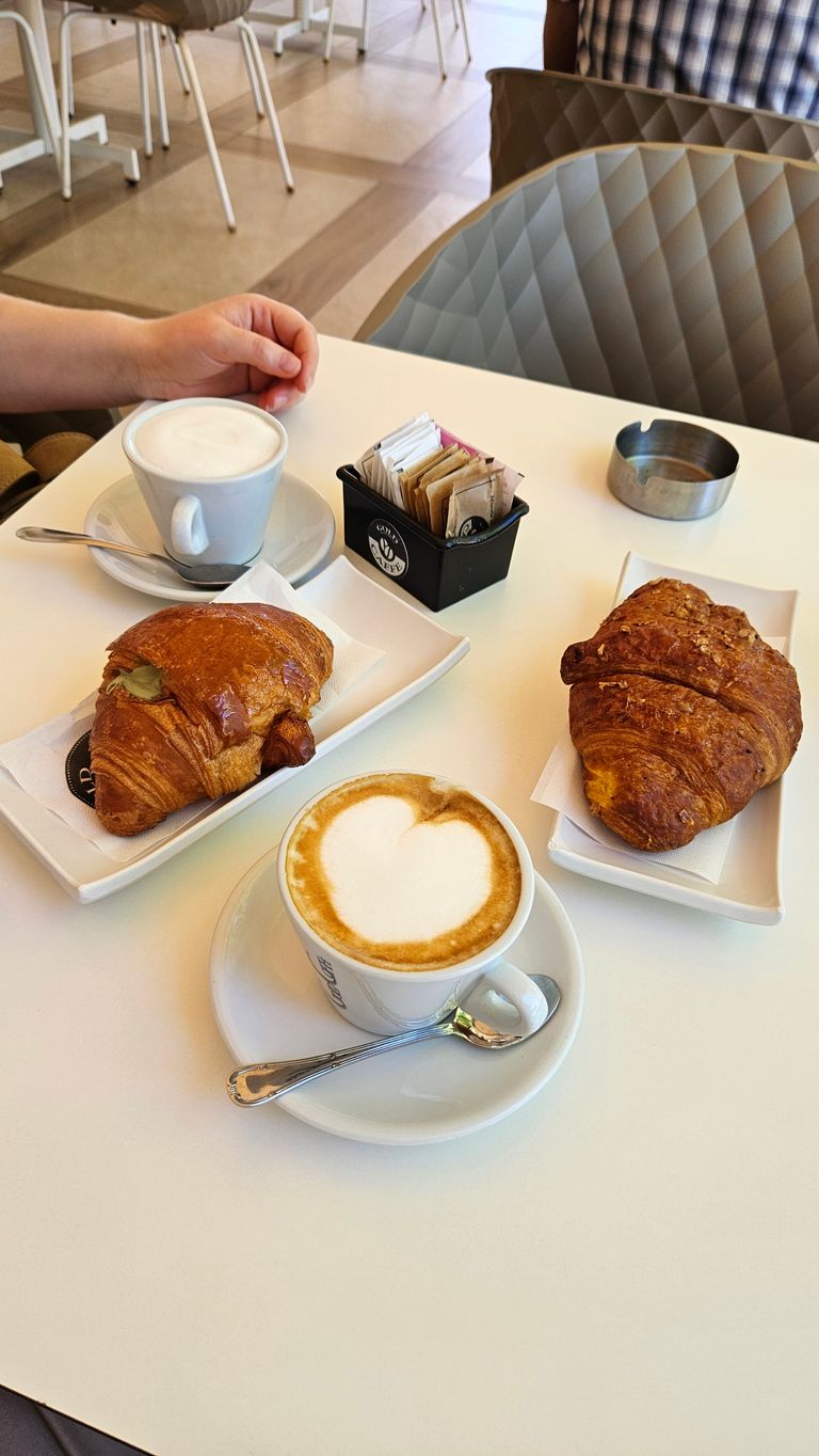 Zwei gefüllte Croissants auf rechteckigen Tellern und zwei Tassen Cappuccino auf einem weißen Tisch in einem Café, mit einer Hand und einem Ablageschälchen im Hintergrund.