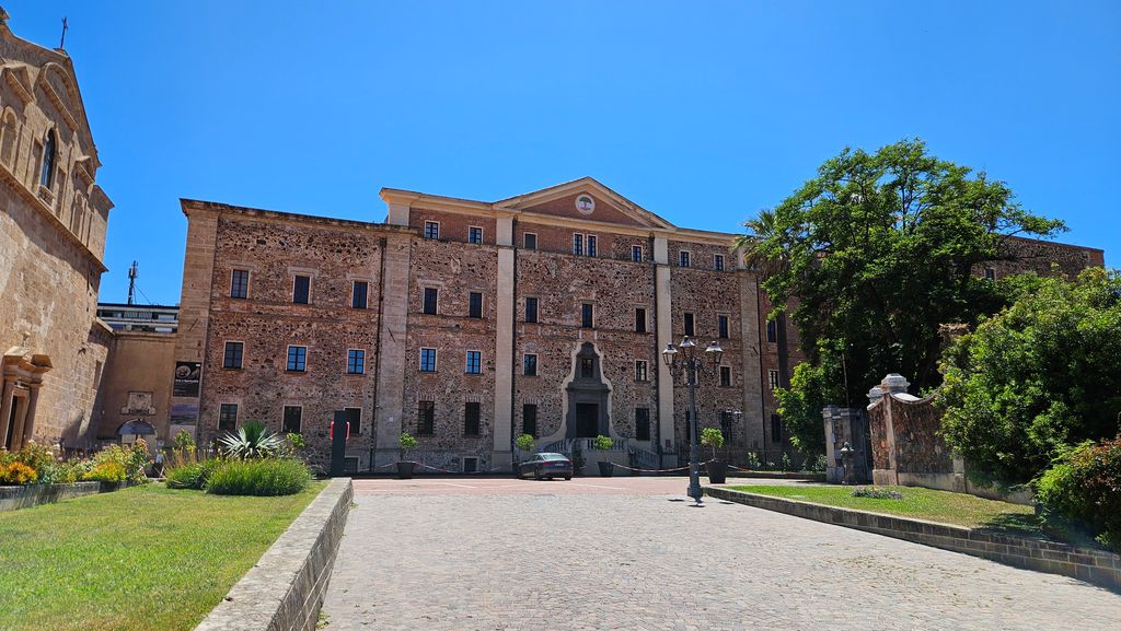 Ein großes, rechteckiges Backsteingebäude mit vielen Fenstern, das an einem sonnigen Tag von Bäumen und einem gepflasterten Weg umgeben ist.