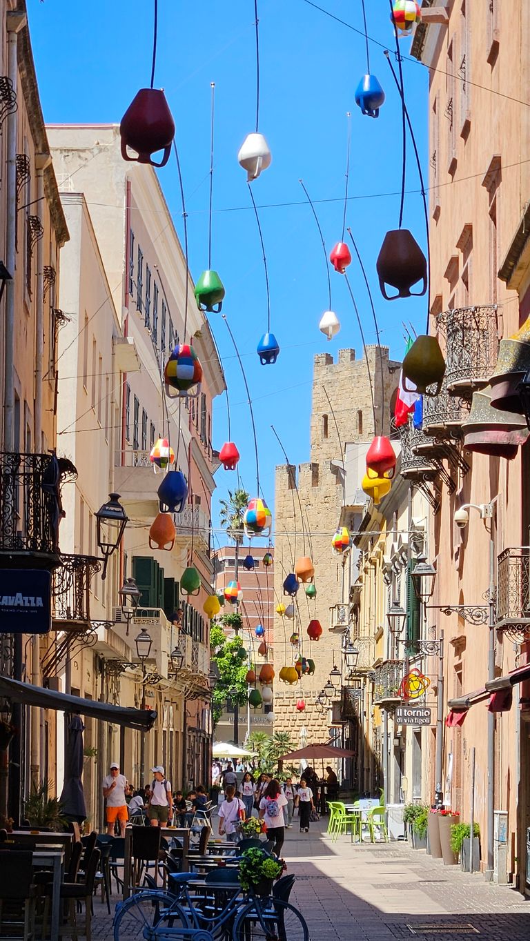 Bunte, hängende Dekorationen über einer belebten Fußgängerzone mit Tischen und Stühlen vor Gebäuden, im Hintergrund eine historische Steinburg unter blauem Himmel.