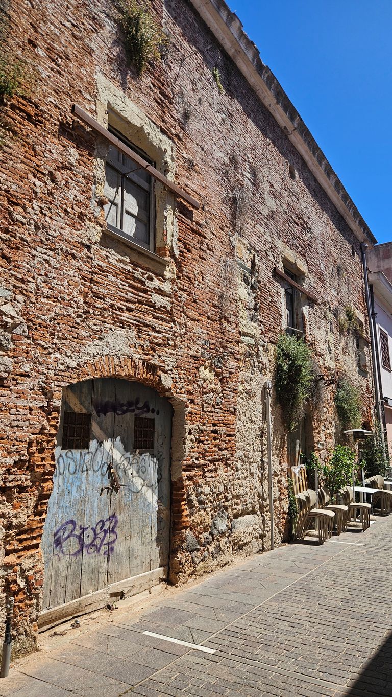 Alte Ziegelsteinwand eines Gebäudes mit bemalter Holztür und zwei Fenstern, die mit Holzlatten gesichert sind. Vor dem Gebäude stehen braune Kunststoffstühle und einige Topfpflanzen auf einem gepflasterten Gehweg unter klarem blauem Himmel.