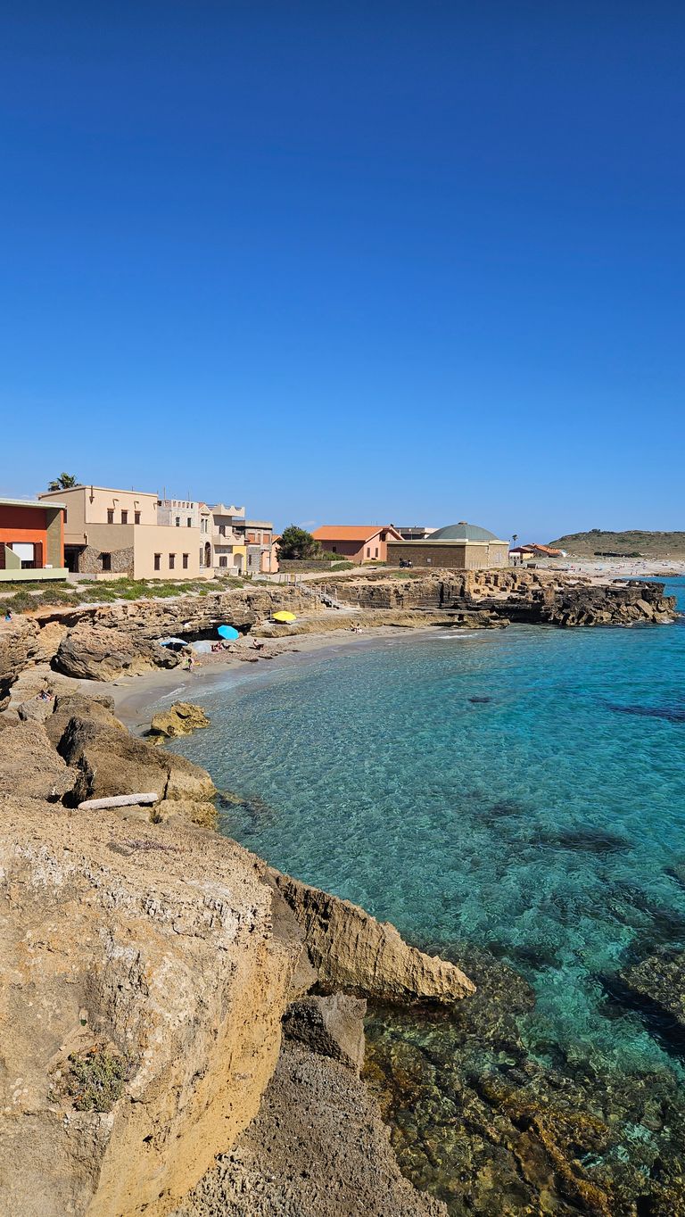 Küstenlandschaft mit klarem, türkisfarbenem Wasser, felsigem Ufer und Häusern am Rand unter einem wolkenlosen blauen Himmel.