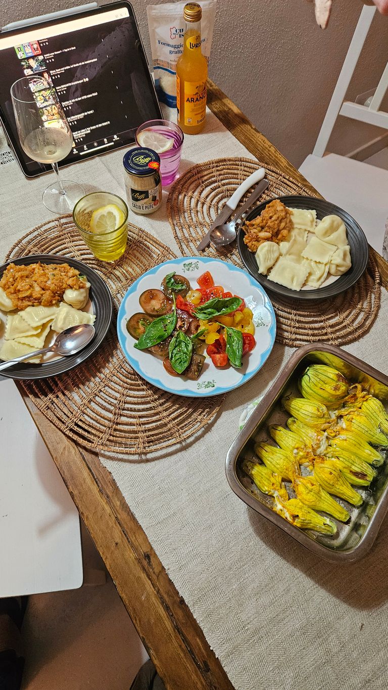 Ein gedeckter Tisch mit zwei Tellern Ravioli, einem Teller mit Tomaten und Basilikum, einem Backblech mit gefüllten Zucchiniblüten sowie zwei Gläsern mit Getränken und einer Flasche Aranciata. Ein Tablet zeigt einen Serien- oder Video-Streaming-Dienst im Hintergrund.